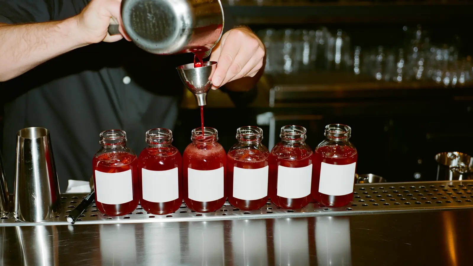 Mains d'un barman transvasant un cocktail pré-mélangé vers des bouteilles en verre sur un plan de préparation