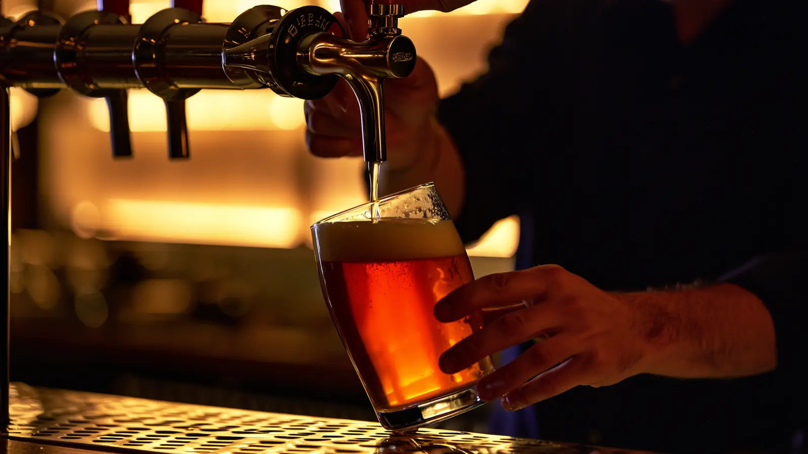 Bartender's hand pulling a draft beer tap handle, glass filling with beer and foam at a dark bar