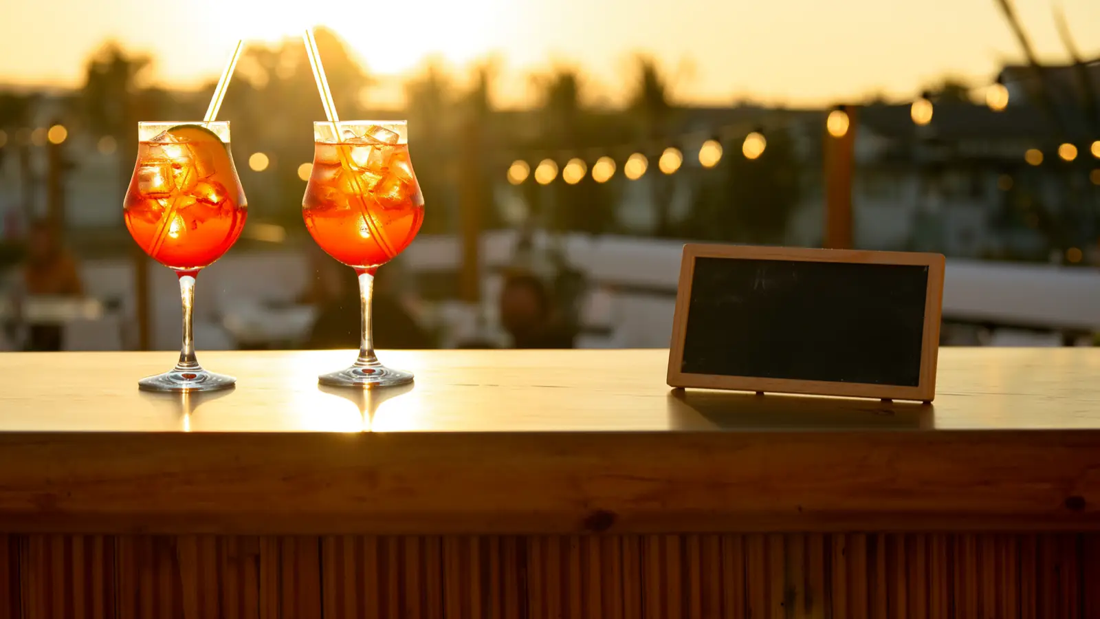 Two spritz cocktails on a golden-lit bar at dusk beside a small blank chalkboard