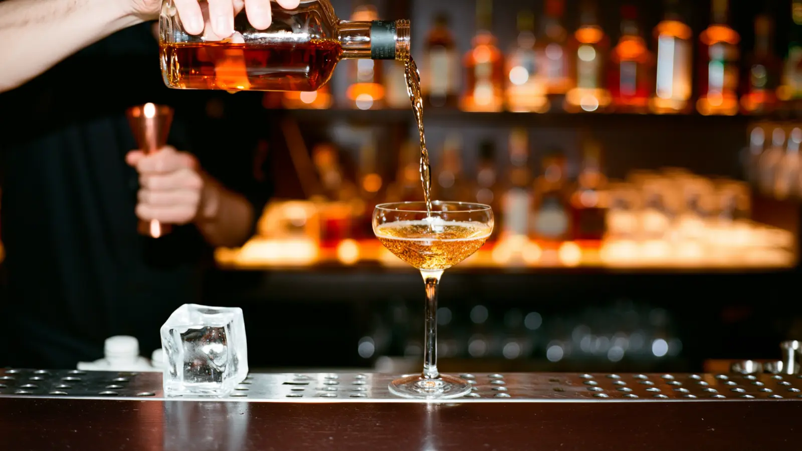 Bartender pouring whiskey into a coupe beside a copper jigger and a clear ice cube at a warmly lit bar