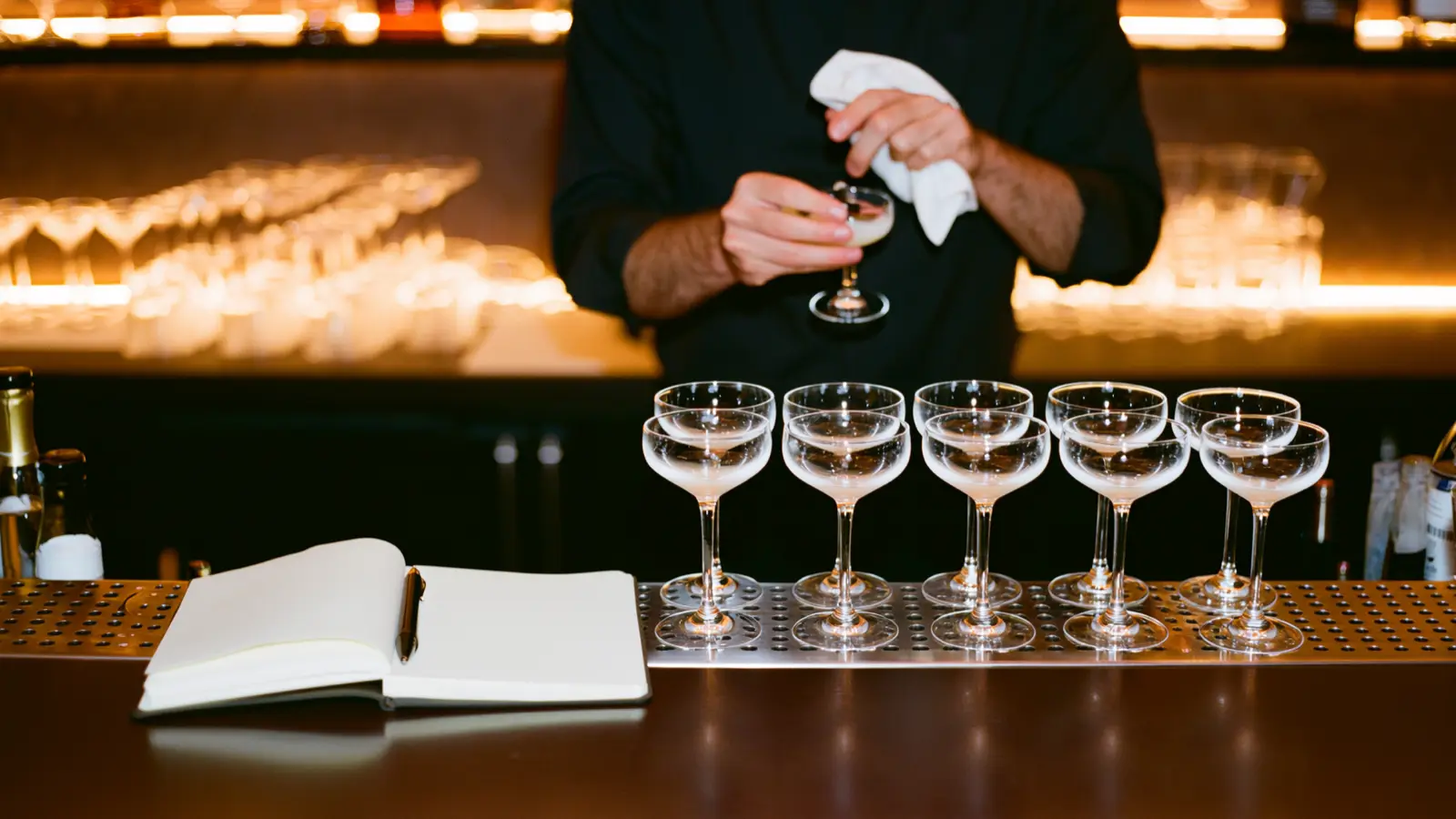 Bartender polishing a coupe behind a row of finished cocktails beside an open training notebook