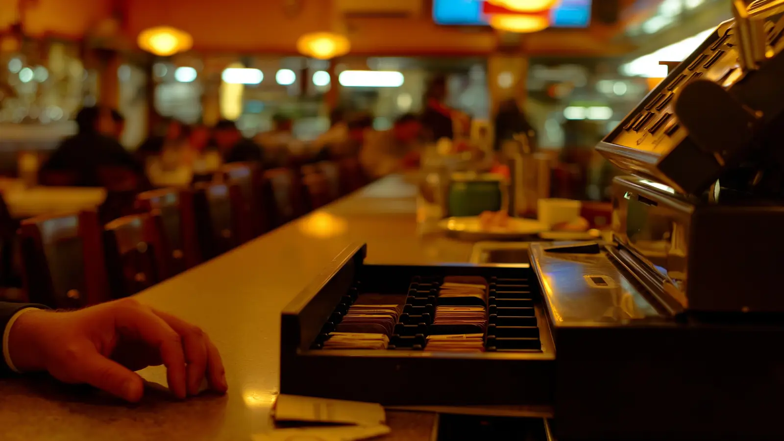 A busy, blurred restaurant dining room behind a nearly empty cash drawer in sharp focus