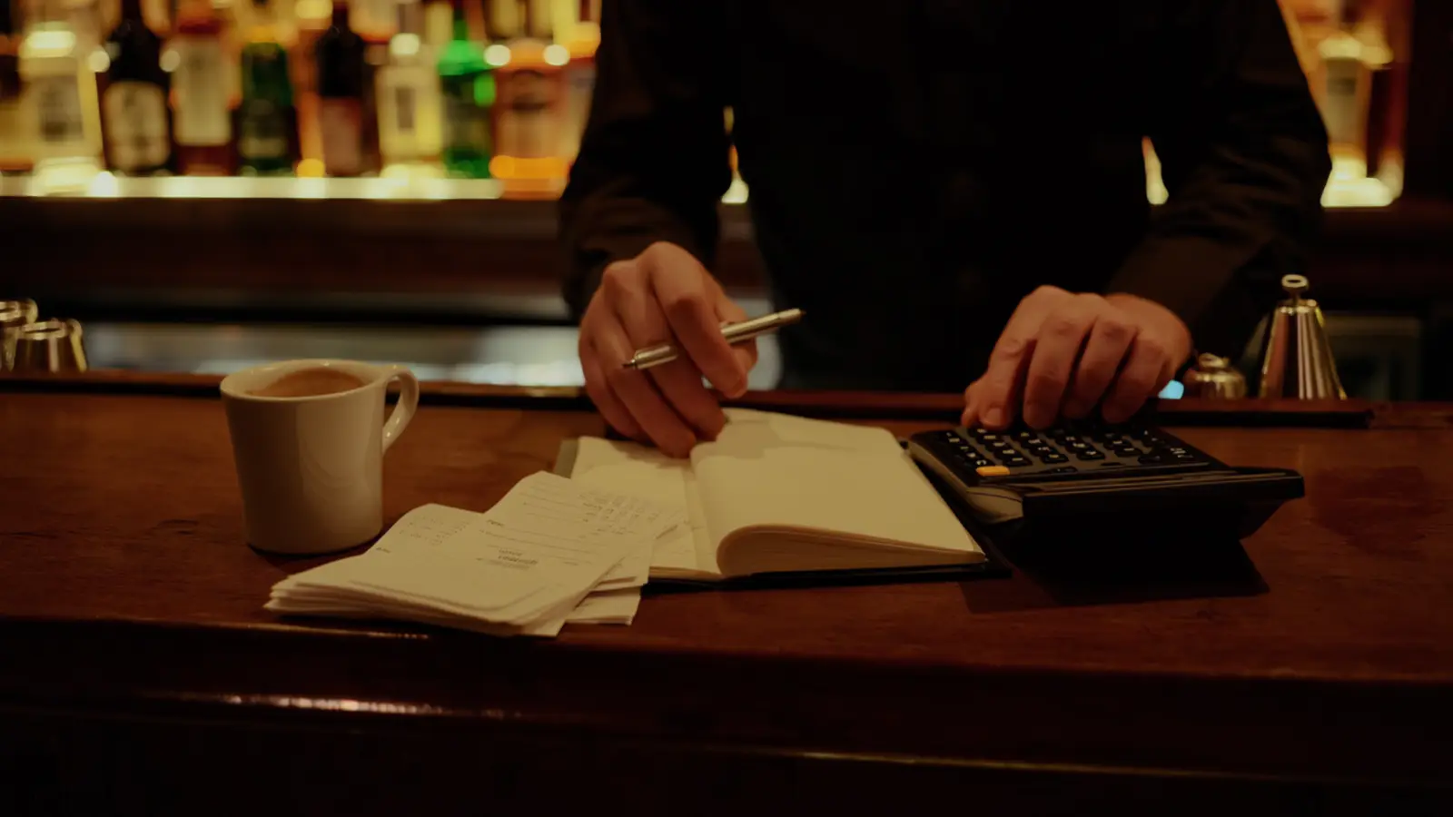 Manager's hands reviewing a ledger beside a calculator and receipts on a bar counter under a warm lamp at close