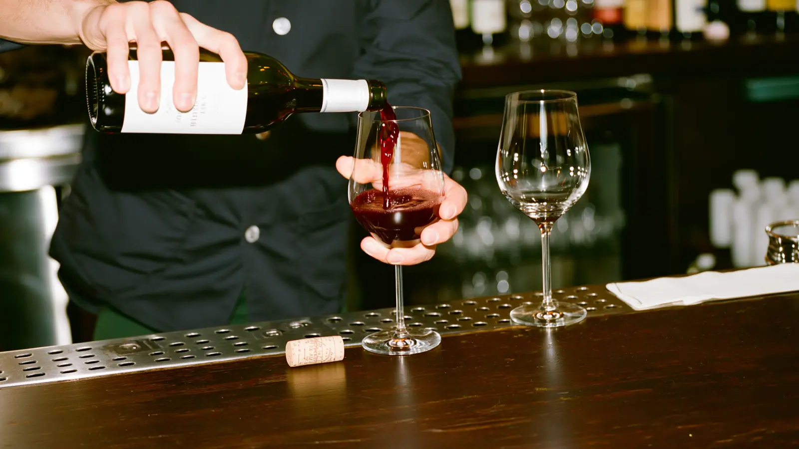 Mains d'un sommelier versant du vin rouge d'une bouteille sans étiquette dans un verre, bar à lumière chaude
